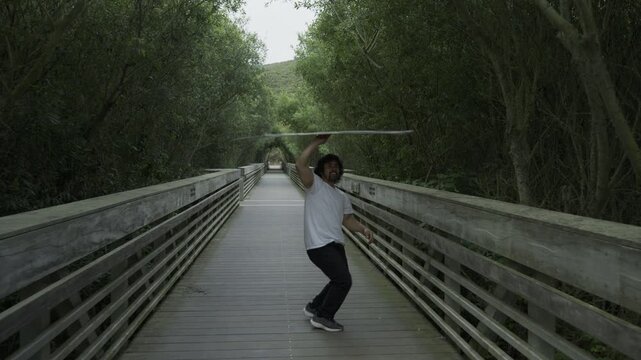 Man Spinning And Balancing Painted Board Overhead On Wooden Footbridge - Steady Cam Shot