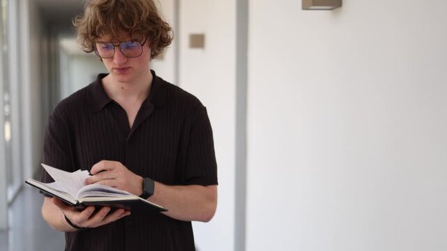 Vertical screen schoolboy reading notes and reviewing notebook in school corridor preparing for upcoming test with focused attention and careful study