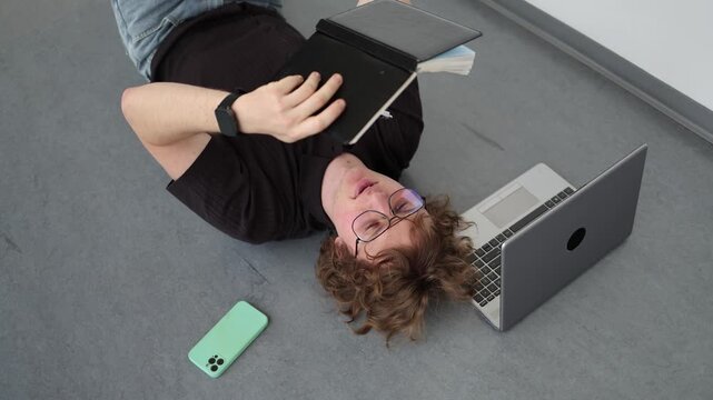 Vertical screen Student studies while lying on floor reading notebook beside laptop reflecting intensive learning long study sessions and dedication to education