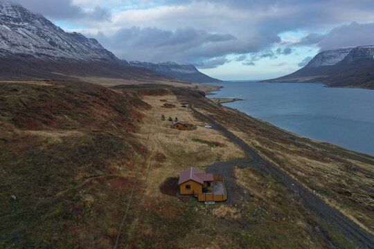 Iceland - 24 June 2026: Aerial view of small wooden cabins situated on a grassy hillside overlooking a calm fjord with snow-capped mountains in the background under a cloudy sky.