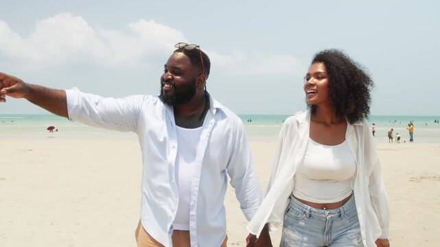 Smiling couple embraces on beach while man points toward distance, enjoying sunny seaside moment together during a relaxing vacation.