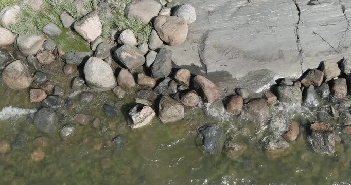 Rocky shoreline or littoral zone, where a massive slab of bedrock meets loose, rounded cobbles and stones in shallow water in summer, aerial view, Uutela, Helsinki, Finland, Europe.
