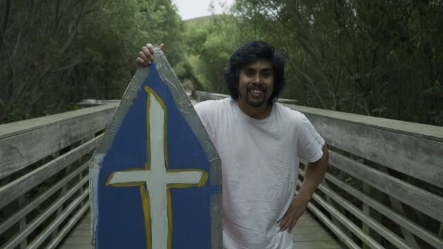 Man Holding Blue Cross Sign on Wooden Footbridge and Smiling - Medium Shot In Slow Motion