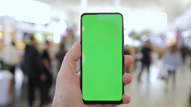 Hand holds phone with green screen in busy public space. Background shows blurred people walking. Ideal for app, social media or advertising mockups. Focus on device, not surroundings