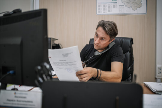 Office worker reading a document while talking on landline phone. Multitasking, heavy workload, and focused business routine.