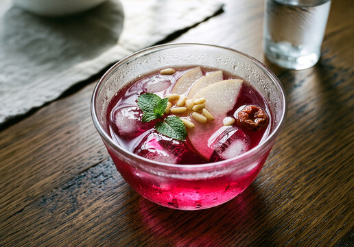Traditional Korean omija fruit punch in transparent glass bowl with sliced Korean pear pine nuts and mint on rustic wooden table
