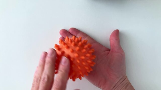A woman rolls an orange massage ball on her palm, doing hand exercises. Top view of a woman's hands on a white background. Stress reduction, relaxation