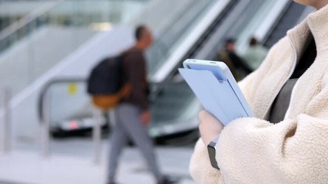 Person holds tablet while standing near moving escalator. Background shows blurred travelers and modern architecture. Focused shot on individual's hands and device
