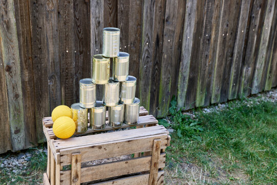 Tin can knockdown game with a pyramid of cans and two yellow balls on a wooden crate outdoors against a fence.