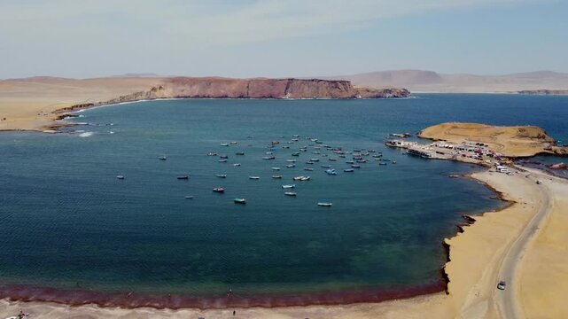 Aerial view of Paracas Bay with numerous small fishing boats anchored in the calm blue water, surrounded by arid desert coastline and sandy beaches in Paracas, Ica, Peru.
