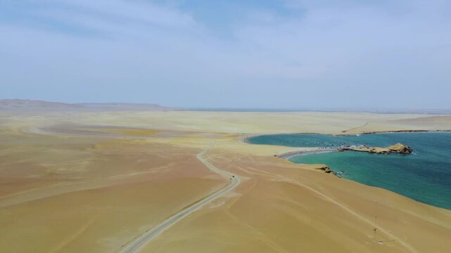 Aerial view of the arid desert landscape and turquoise coastline of Paracas National Reserve, with a winding road cutting through the sand in Paracas, Ica, Peru.