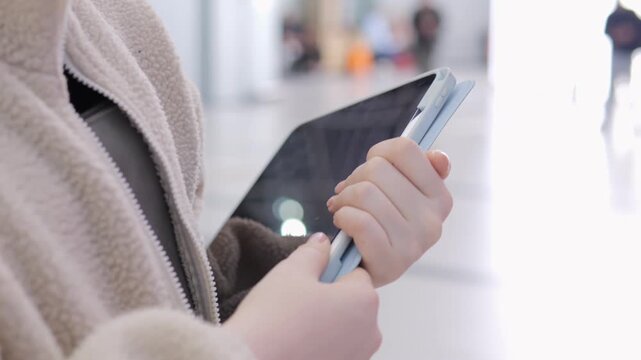 Close-up of hands holding a tablet and stylus. Person wears light beige fleece jacket. Background shows blurred indoor space with people moving. Focus stays on device and user's interaction