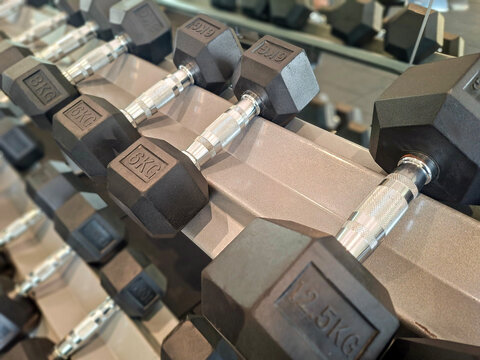 row of black rubber-coated hexagonal dumbbells with chrome knurled handles neatly organized on a metallic storage rack in a commercial gym.