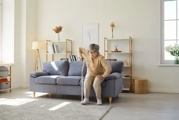 Fototapeten Musik Portrait a of senior gray-haired elderly injured woman getting up from the couch with the help of her crutch in living room at home. Mature female person using walking cane indoors.  © Studio Romantic