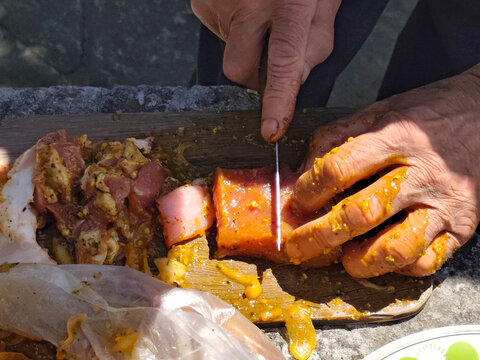 Close-up of hands preparing marinated meat for grilling outdoors. Pieces of raw meat coated with seasoning and marinade are being cut and threaded onto metal skewers. Plastic bag with marinated meat, 