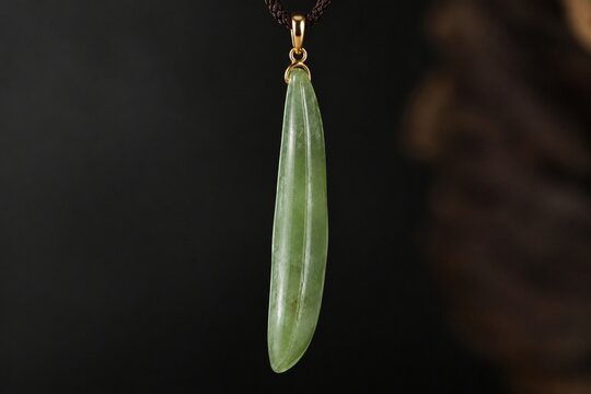 A slender green jade pendant shaped like a silique on a dark background.