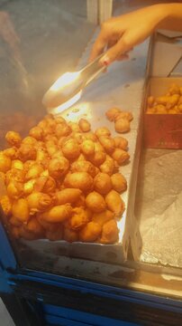 Close-up of a street food vendor's hand using metal tongs to pick up mini molen (Indonesian fried banana pastry) from a glass display, representing traditional Asian snacks and local culinary business
