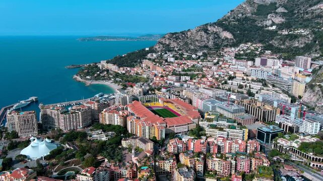 Aerial orbit over Stade Louis II football stadium in the Fontvieille district of Monaco, with the port, apartment buildings, coastal cliffs and the Mediterranean Sea on the French Riviera.