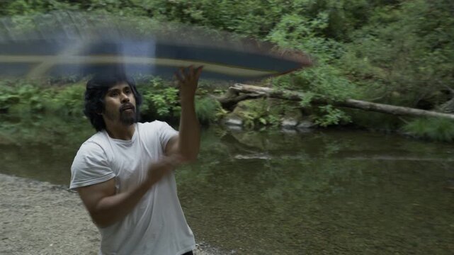 Man Spinning Painted Board on Gravel Riverbank Among Trees - Medium Steady Cam Shot