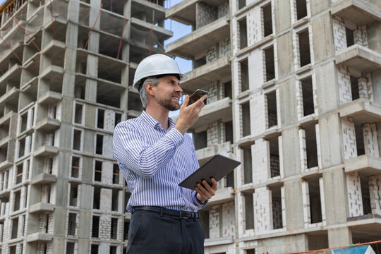 Side view of male engineer standing against background of buildings under construction working in construction helmet