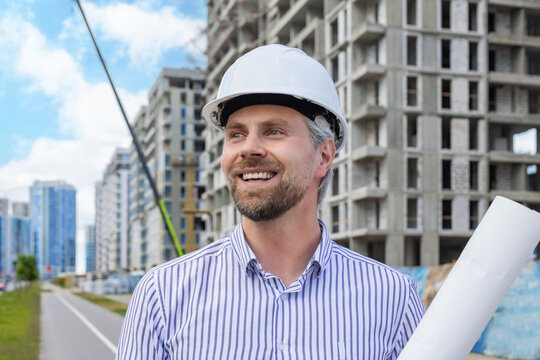 Close-up portrait of a young smiling man architect wearing a helmet with blueprint plans looking at the camera in building under construction