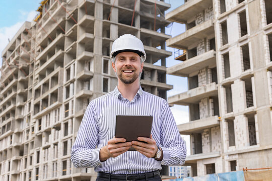 Handsome man with worker's helmet holding digital tablet while looking at camera, background buildings under construction.