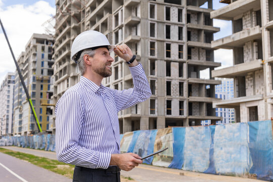 Side view of male engineer standing against background of buildings under construction working in construction helmet