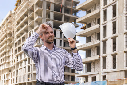 Tired male engineer taking off safety helmet touching forehead while working at construction site