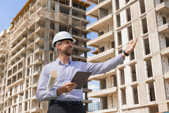 Side view of male engineer standing against background of buildings under construction working in construction helmet