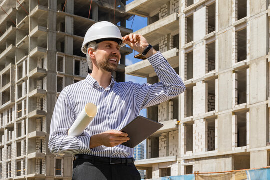 Side view of male engineer standing against background of buildings under construction working in construction helmet
