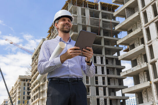 Male engineer standing against background of buildings under construction working in construction helmet
