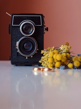 Vintage twin-lens reflex camera, gold wedding rings, and Sardinian wildflowers on a white table with pink wall background