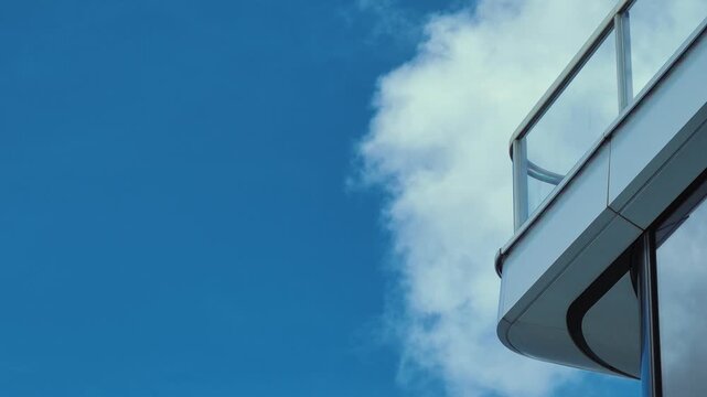 A sleek modern building's balcony against a clear blue sky with a single fluffy white cloud moving from left to center