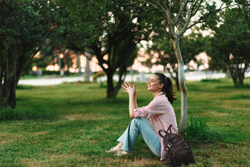 A reflective woman sits on grass in a park, hands clasped, in contemplation as trees and soft light...