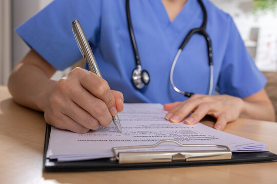 Healthcare doctor clipboard writing medical form stethoscope scrub patient document, clinician writing paper form during consultation clinic with stethoscope, blue scrub, hand, pen, desk