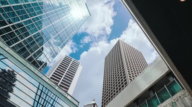 A low-angle shot of towering skyscrapers in downtown Hong Kong, reflecting the blue sky and fluffy white clouds