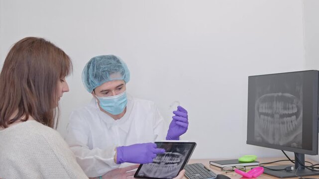 orthodontist in his office discussing a teeth straightening treatment plan with a female patient, showing a digital x-ray on a tablet and explaining how invisible aligners work
