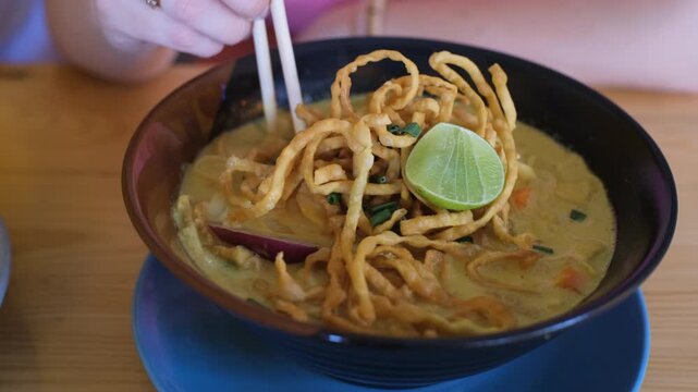 Close-up of a young woman stirring a bowl of traditional northern Thai Khao Soi curry noodle dish before eating. Thai food concept