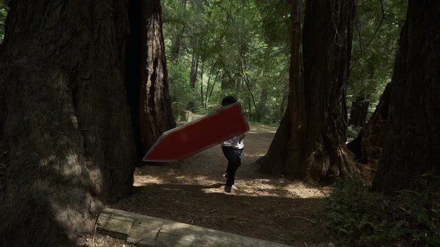 Man Carrying Red Board Through Giant Tree Trunks Forest And Doing Professional Tricks