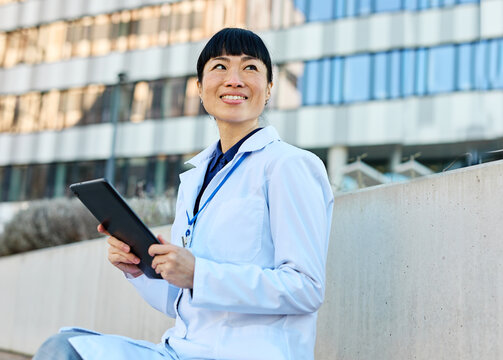 Portrait of an asian female woman doctor or nurse and  health care worker holding a cup of coffee and using a tablet computer in front of hospital or clinic outside