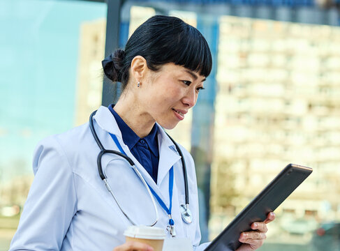 Portrait of an asian female woman doctor or nurse and  health care worker holding a cup of coffee and using a tablet computer in front of hospital or clinic outside