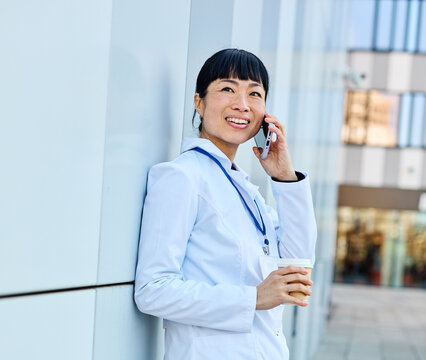 Portrait of an asian female woman doctor or nurse and  health care worker using a smartphone mobile phone holding a cup of coffee   in front of hospital or clinic outside