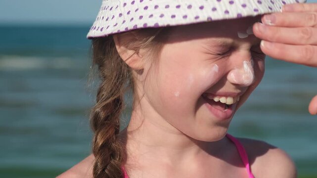 Close up mother applying protective sunscreen on daughter at beach during summer vacation. Female hand putting sun lotion on child face. Happy little girl with sunblock taking sunbath on sea coast