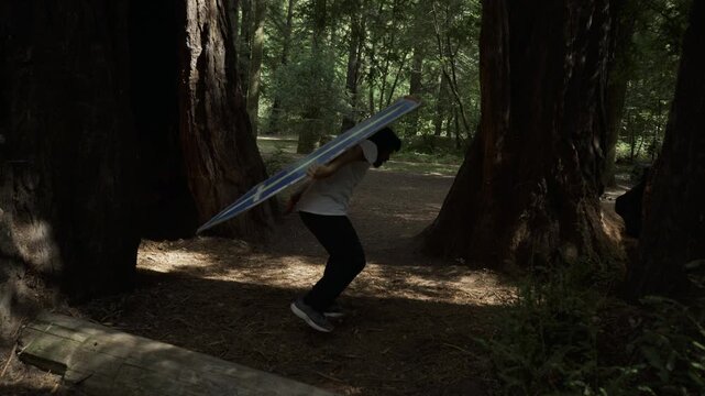 Man Bending Low While Swinging Painted Board Among Redwood Trunks - Tilt Shot
