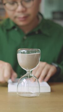 Closeup Turning Hourglass Upside Down to Continue Studying and Reading Book on Wooden Desk.