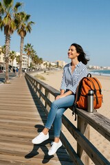 Fototapeta premium Young woman sits on wooden boardwalk railing. Orange backpack and thermos by her side. Blue sky, sand beach, ocean waves, palm trees visible. She smiles with eyes closed enjoying sunny day.