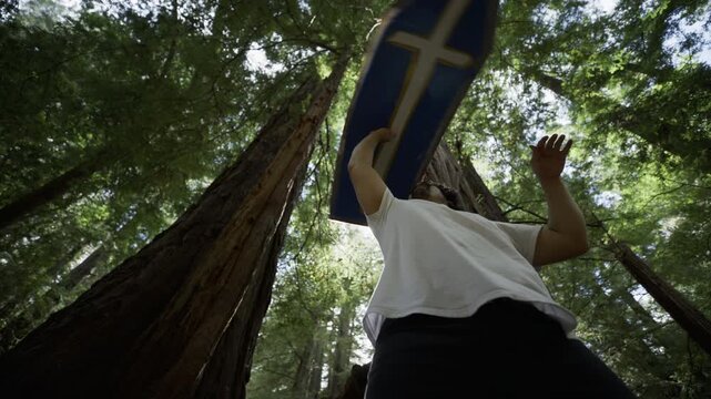 Slow Motion Low Angle Of Man Spinning And Balancing Blue Cross Sign Overhead Among Redwood Trunks