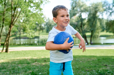 Obraz premium A cheerful little boy holds a blue ball while playing in a summer park.