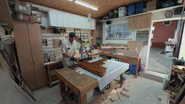 Industrious workshop owner wearing white overalls coating raw pine timber boards with rich brown wood stain using small hand roller in authentic private manufacturing carpentry studio wide angle shot
