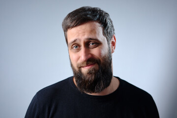Naklejka premium Bearded man gives a wry, friendly smile in a casual headshot against a neutral gray background. Natural lighting highlights his expressive eyes and textured facial hair
